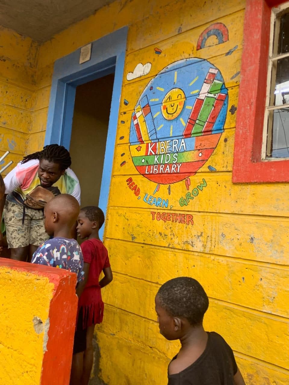 The yellow front of the Kibera Kids Library, painted with a sun and books and the words read, learn, grow together