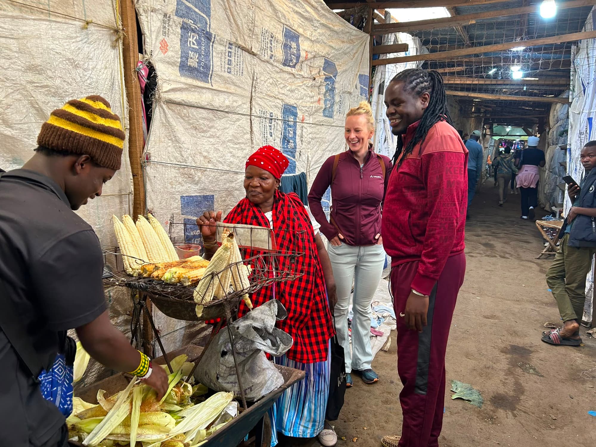 A vendor grilling maize at her stall in the market while Sammy and a guest look on