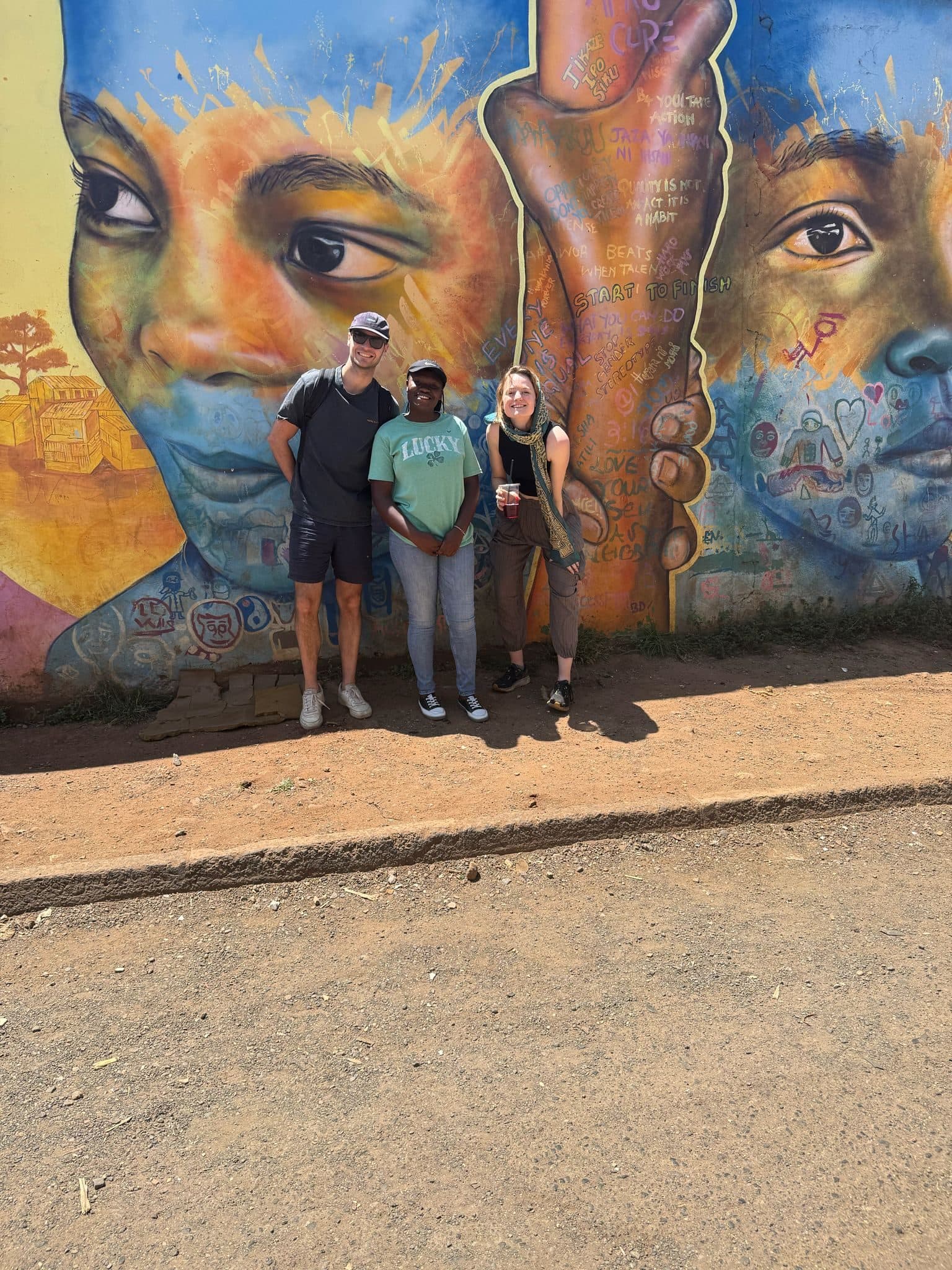 Three guests standing in front of a large painted mural of two faces on a Kibera wall
